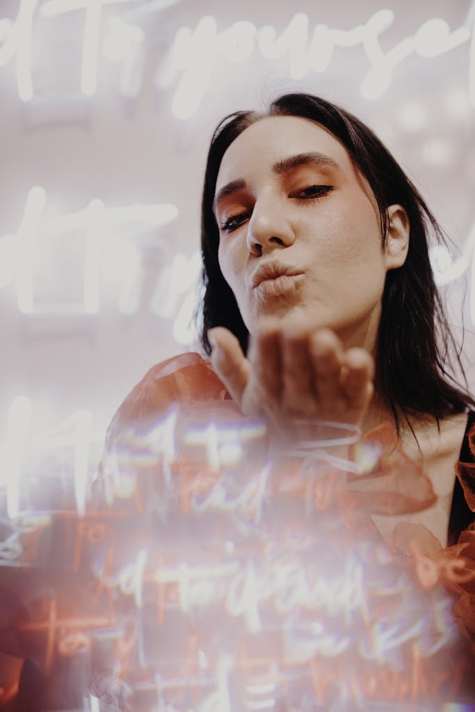 Woman in red makeup blowing a kiss, surrounded by neon lights creating a romantic and vibrant atmosphere.