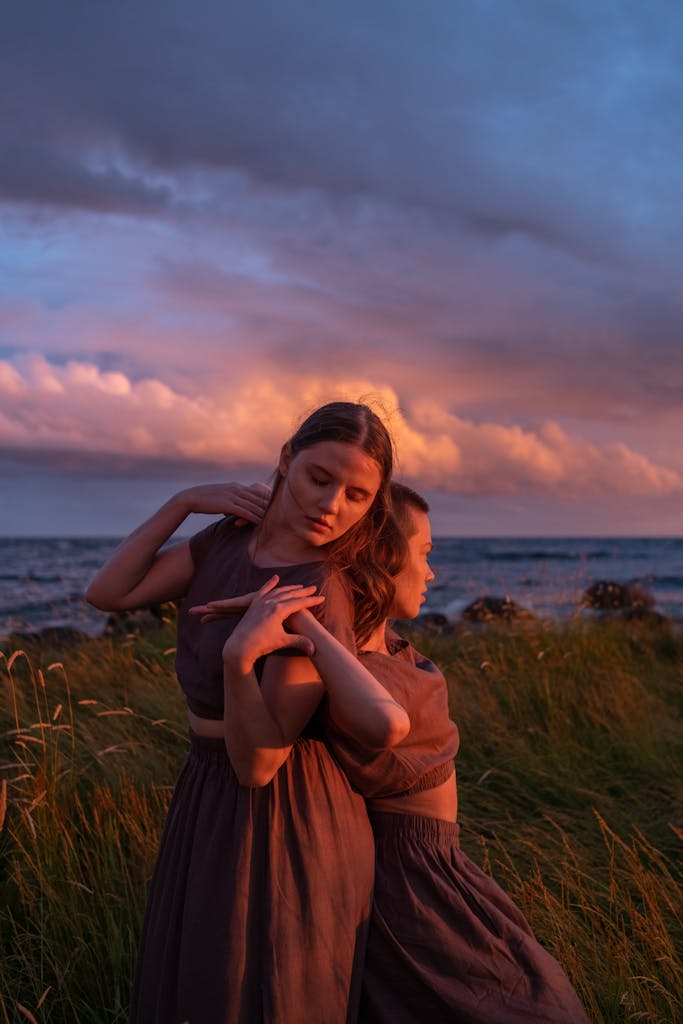 Two dancers in flowing dresses gracefully pose back-to-back amid a stunning sunset by the seaside.